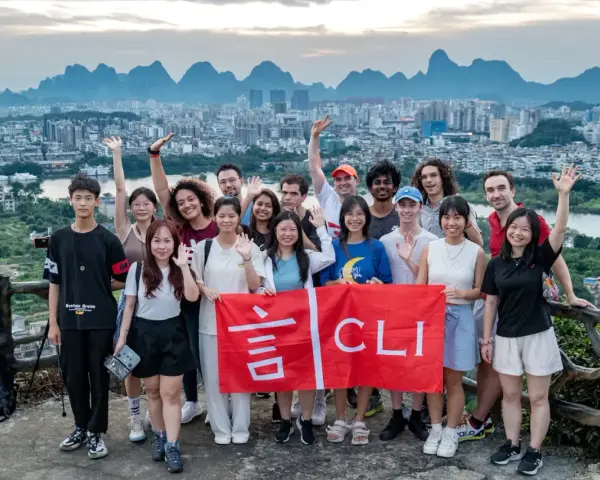 Grupo de jóvenes sonríe y saluda, sosteniendo una bandera roja con caracteres chinos y "CLI". Un paisaje urbano y montañoso de China al fondo, ideal para viajar a China por libre.