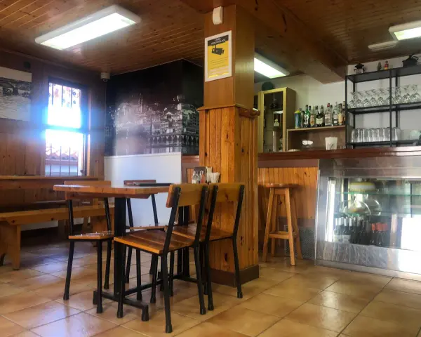 Mesas de madera y sillas listas para comer en Pontedeume. Barra con botellas y vasos, y un mural de la ciudad.