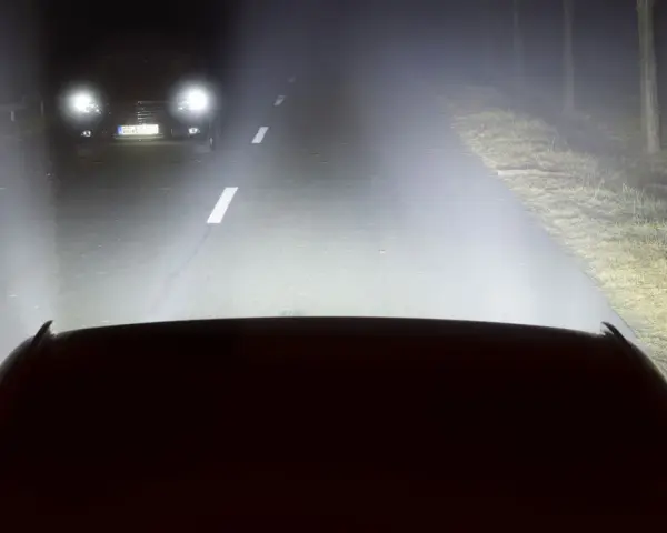 Luces de carretera o largas iluminan la noche, revelando un coche adelante y el camino.