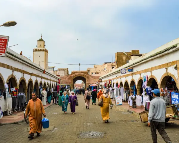 Mercado vibrante en Essaouira que ver: gente comprando ropa y artesanías bajo arcos blancos, con una mezquita al fondo.