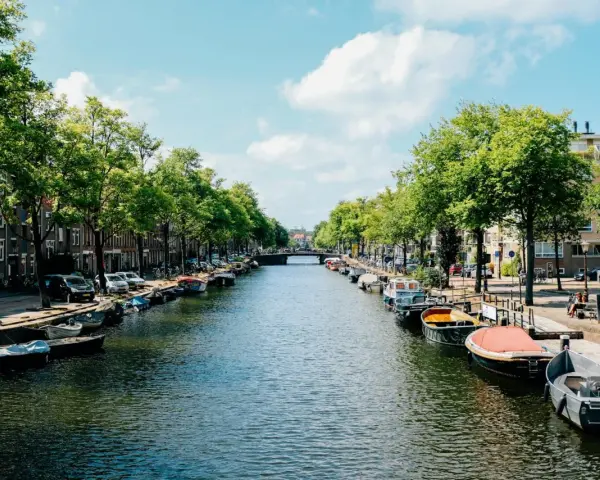 Canal de Ámsterdam con barcos, árboles y edificios bajo un cielo azul con nubes.