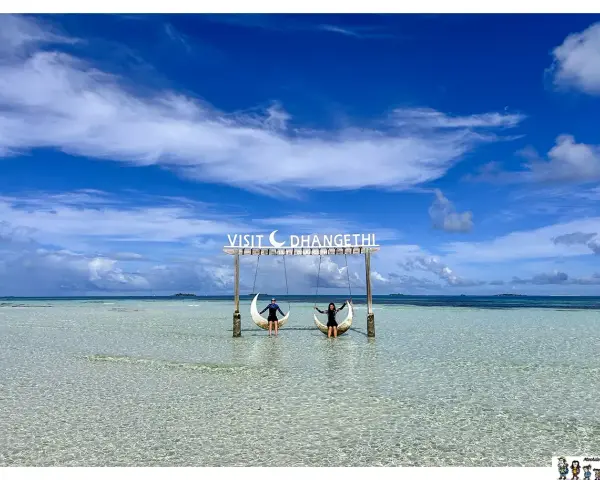 Dos personas disfrutan de columpios en forma de luna sobre aguas cristalinas. Un cartel dice "Visit Dhangethi", mostrando que las Maldivas low cost son posibles.