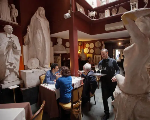 Un camarero sirve copas de vino en un restaurante único, rodeado de esculturas. Un lugar ideal donde comer en Roma.