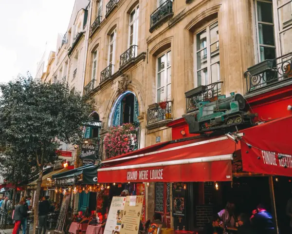 Calle parisina con restaurantes y mesas al aire libre, ideal para comer barato en París. Un tren decorativo adorna un toldo rojo.