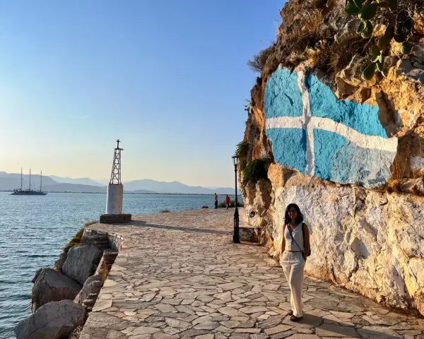Una mujer posa junto a una roca con la bandera griega pintada. Un faro y un velero adornan la costa, ideal para saber qué ver en Grecia en una semana.