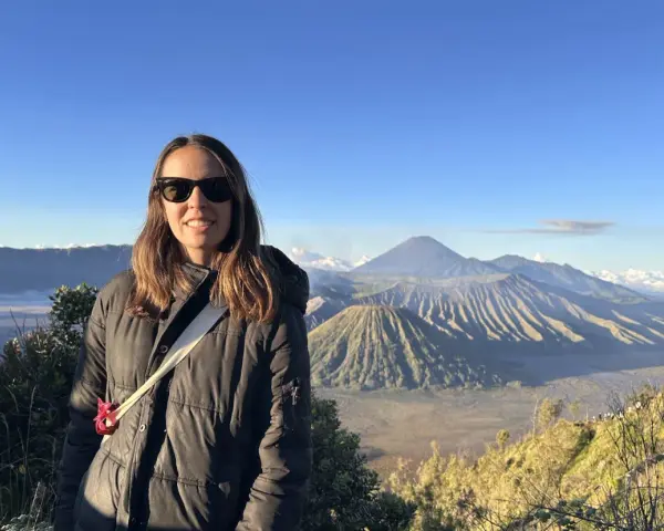 Mujer sonríe con gafas de sol frente a un paisaje volcánico en su ruta por Indonesia.