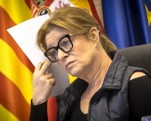 Mujer con gafas y chaleco oscuro, con banderas de España y Europa al fondo. Planeando su próximo viaje por España.