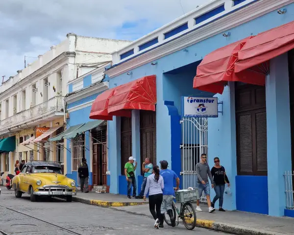 Gente paseando por una calle cubana, con un coche clásico amarillo y edificios de colores. Ideal para viajar a Cuba por libre.