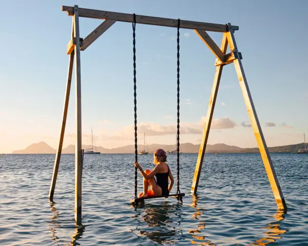 Mujer en columpio sobre el mar, ideal para saber donde viajar en febrero. Paisaje idílico con veleros y montañas al atardecer.