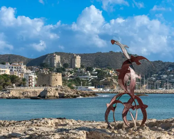 Escultura de aves metálicas en primer plano, con el castillo de Moraira y el puerto al fondo. Descubre qué ver en Moraira.
