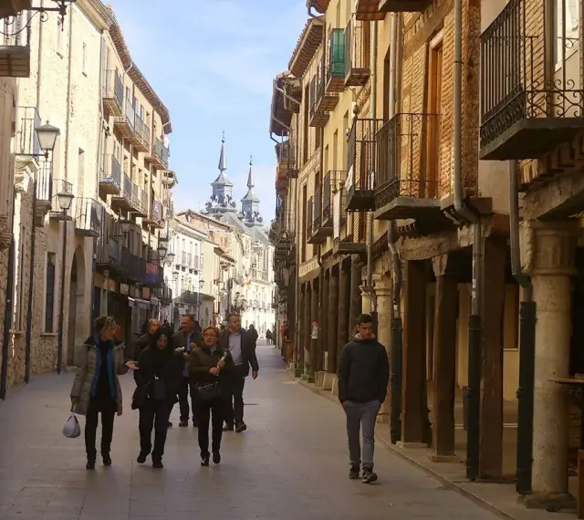 Gente pasea por una calle porticada, un lugar ideal para descubrir qué ver en Burgo de Osma. Edificios históricos y torres se alzan al fondo.