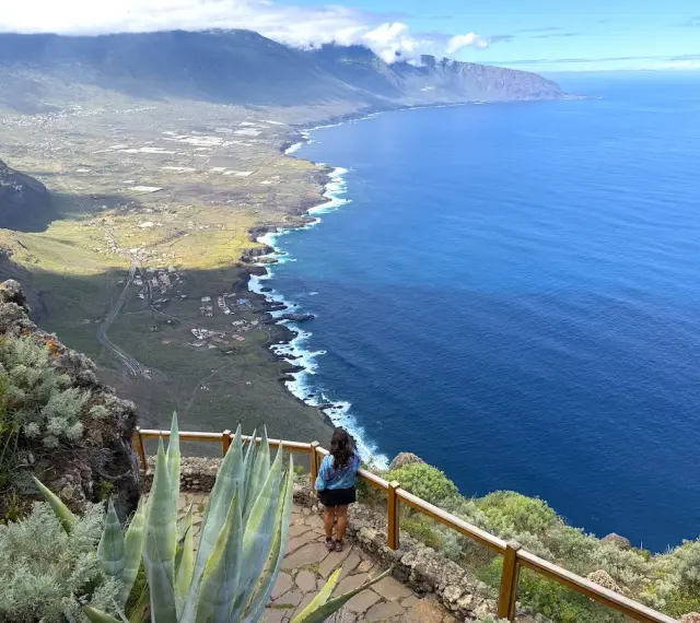 Mujer admira la costa de El Hierro, con el mar azul intenso y montañas verdes.
