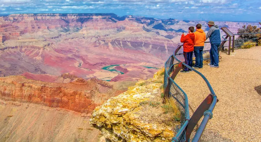 Tres personas admiran la inmensidad del cañón del Colorado, con sus capas rocosas de tonos rojizos y un río turquesa serpenteando en el fondo.