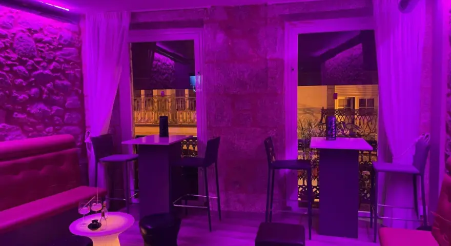 Ambiente moderno y acogedor para comer en Coruña. Mesas altas, taburetes y sofás rojos bajo luces violetas.