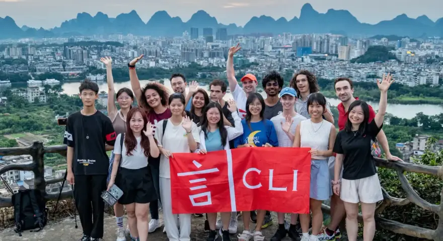 Grupo de jóvenes sonríe y saluda, sosteniendo una bandera roja con caracteres chinos y "CLI". Un paisaje urbano y montañoso de China al fondo, ideal para viajar a China por libre.