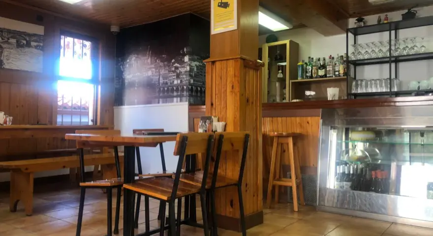 Mesas de madera y sillas listas para comer en Pontedeume. Barra con botellas y vasos, y un mural de la ciudad.