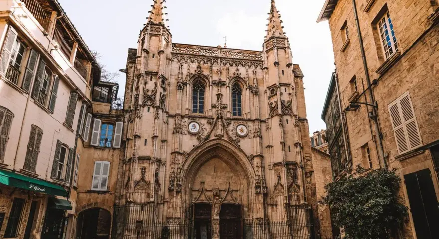 Fachada de una iglesia gótica con detalles ornamentados y dos torres. A los lados, edificios antiguos de piedra y fachadas de color claro. Un lugar que ver en Avignon.