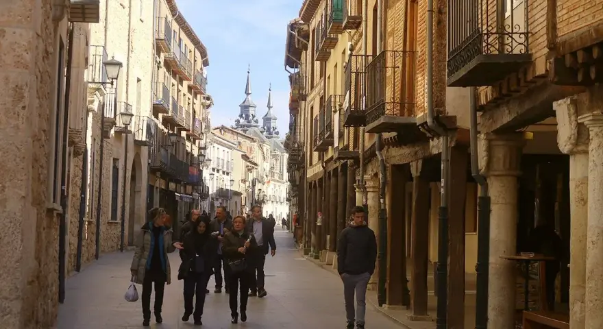 Gente pasea por una calle porticada, un lugar ideal para descubrir qué ver en Burgo de Osma. Edificios históricos y torres se alzan al fondo.
