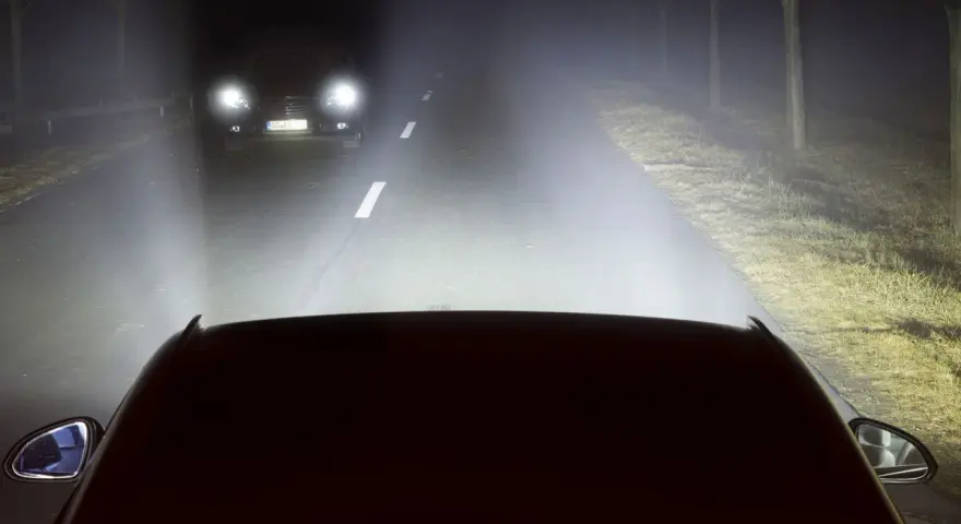 Luces de carretera o largas iluminan la noche, revelando un coche adelante y el camino.