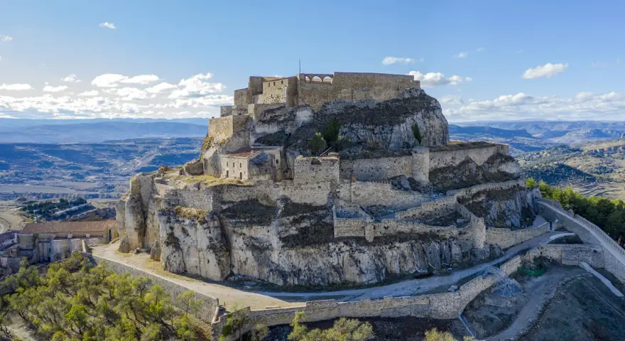 Castillo de Morella sobre un peñón rocoso, con murallas y edificios de piedra bajo un cielo azul con nubes.