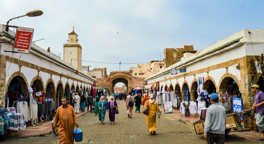 Mercado vibrante en Essaouira que ver: gente comprando ropa y artesanías bajo arcos blancos, con una mezquita al fondo.