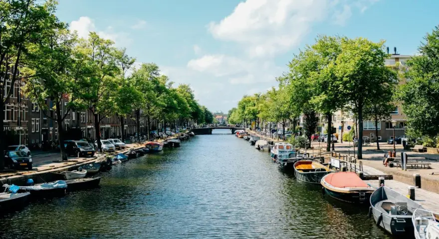 Canal de Ámsterdam con barcos, árboles y edificios bajo un cielo azul con nubes.