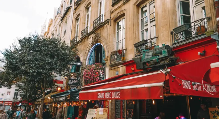 Calle parisina con restaurantes y mesas al aire libre, ideal para comer barato en París. Un tren decorativo adorna un toldo rojo.