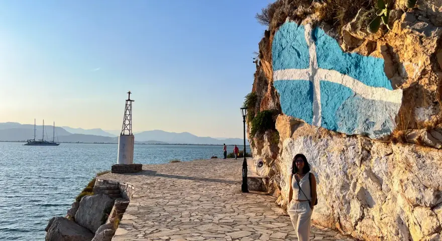 Una mujer posa junto a una roca con la bandera griega pintada. Un faro y un velero adornan la costa, ideal para saber qué ver en Grecia en una semana.