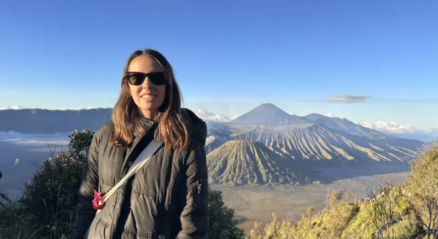 Mujer sonríe con gafas de sol frente a un paisaje volcánico en su ruta por Indonesia.