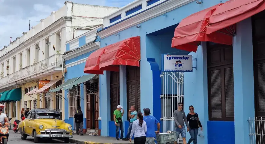 Gente paseando por una calle cubana, con un coche clásico amarillo y edificios de colores. Ideal para viajar a Cuba por libre.