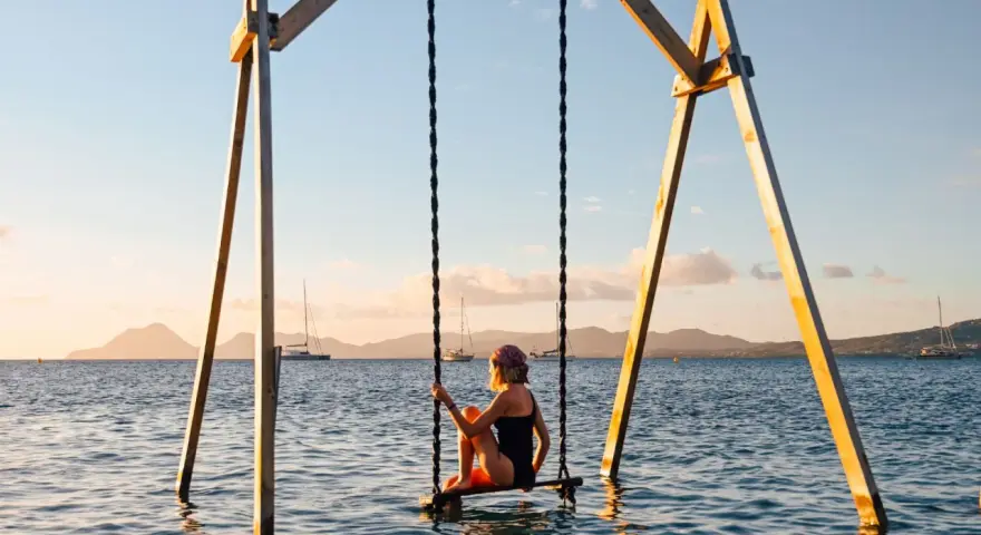Mujer en columpio sobre el mar, ideal para saber donde viajar en febrero. Paisaje idílico con veleros y montañas al atardecer.
