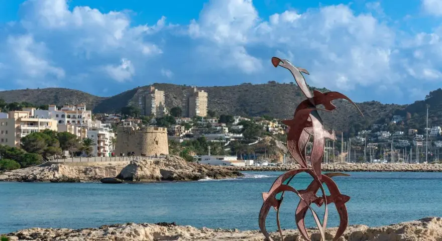 Escultura de aves metálicas en primer plano, con el castillo de Moraira y el puerto al fondo. Descubre qué ver en Moraira.
