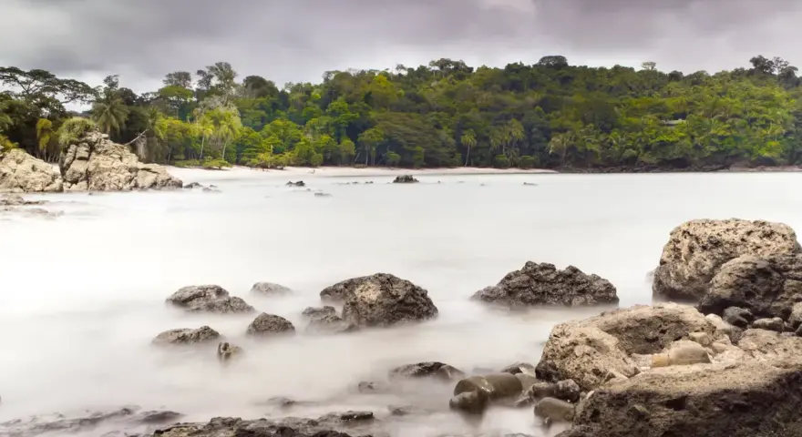 Costa Rica por libre: rocas y mar embravecido en una playa tropical, con densa vegetación al fondo.