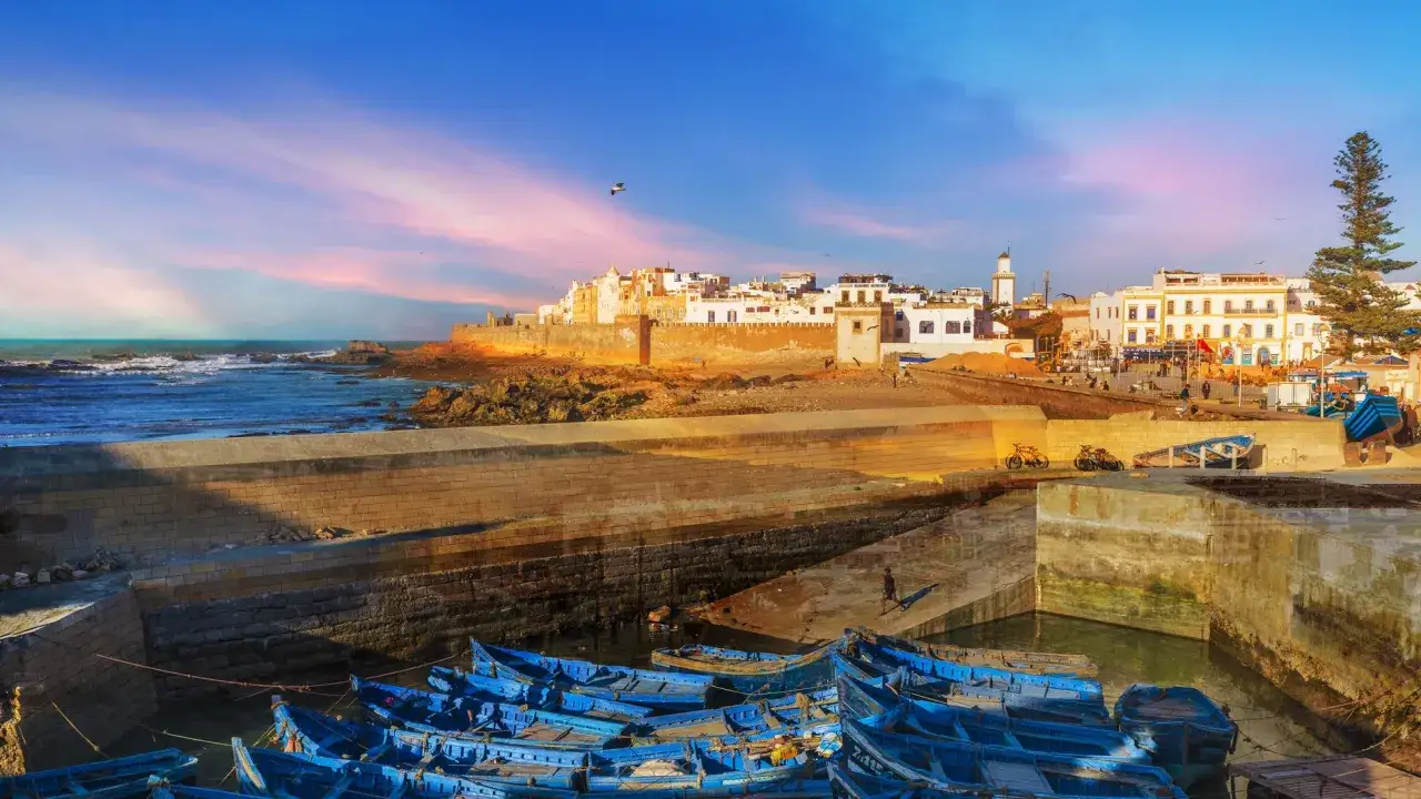 Atardecer en Essaouira: un puerto con barcas azules, murallas doradas y casas blancas bajo un cielo rosado. &iexcl;Essaouira que ver!