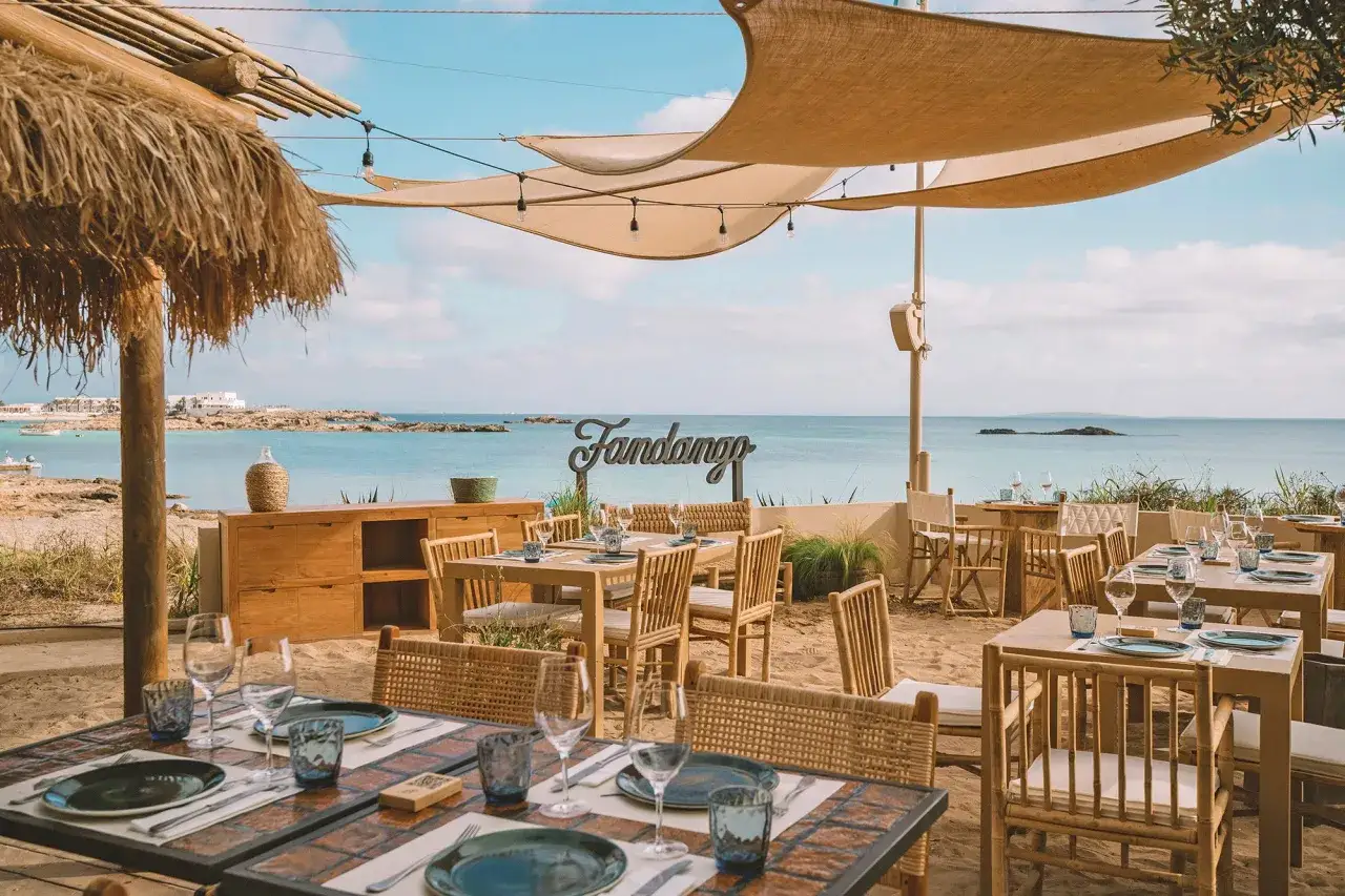 Mesas de restaurante en la arena con vistas al mar. Un lugar ideal donde comer en Formentera, con el cartel 