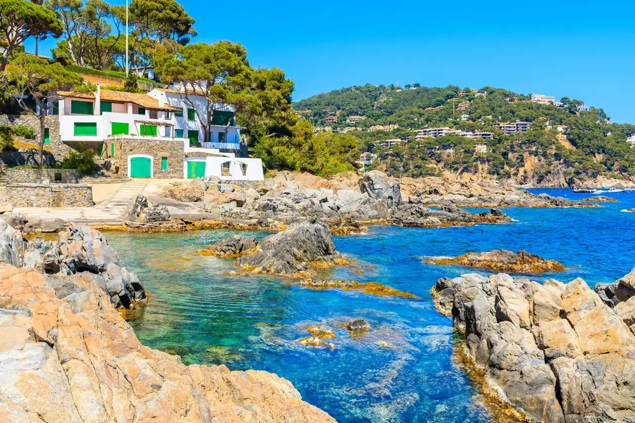 Agua turquesa y rocas doradas en Calella de Palafrugell, con casas blancas y verdes en la ladera.