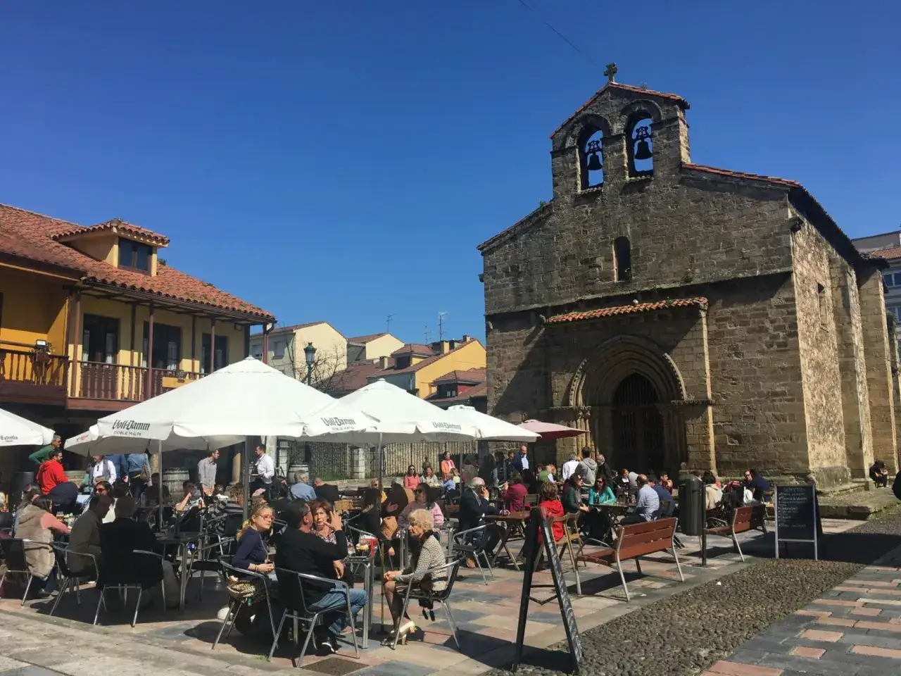 Gente disfrutando de un d&iacute;a soleado para comer en Avil&eacute;s, con mesas al aire libre frente a una iglesia antigua.