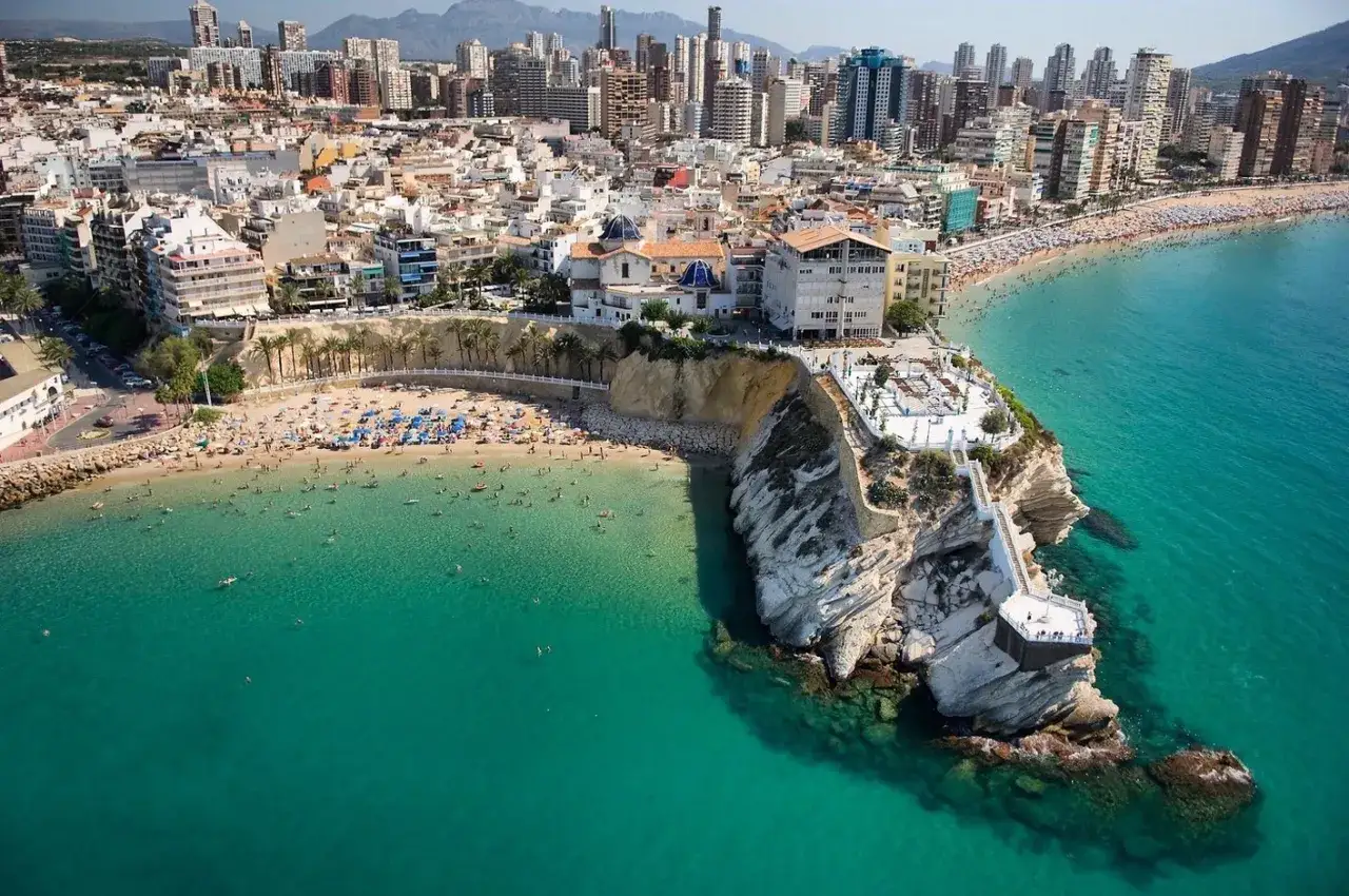 Playa con gente disfrutando del sol y el mar turquesa. Un acantilado blanco con escaleras y un mirador. Ideal para saber donde viajar en julio barato.