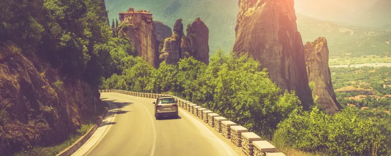 Coche recorre carretera escénica hacia Meteora, Grecia. Monasterio en acantilado y formaciones rocosas imponentes. ¡Un viaje inolvidable a Grecia!