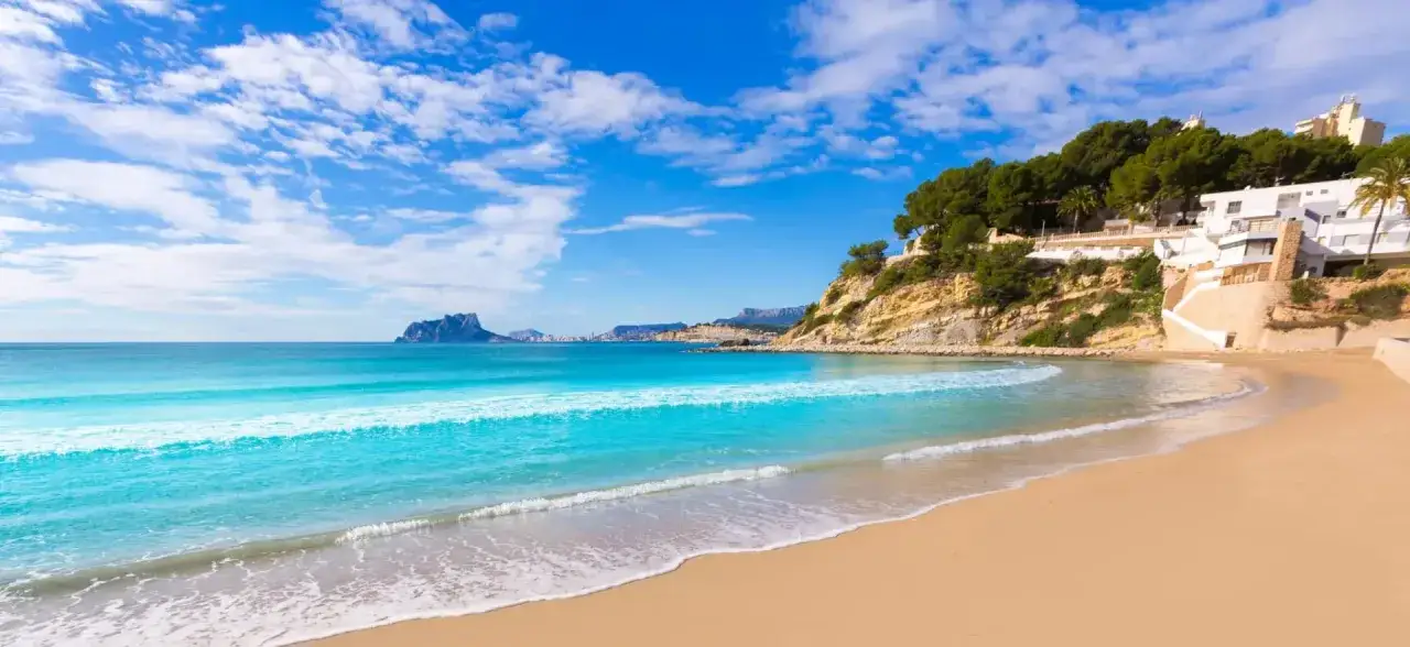 Playa de arena dorada y aguas turquesas, ideal para descubrir qu&eacute; ver en Moraira. Edificios blancos coronan el acantilado verde.