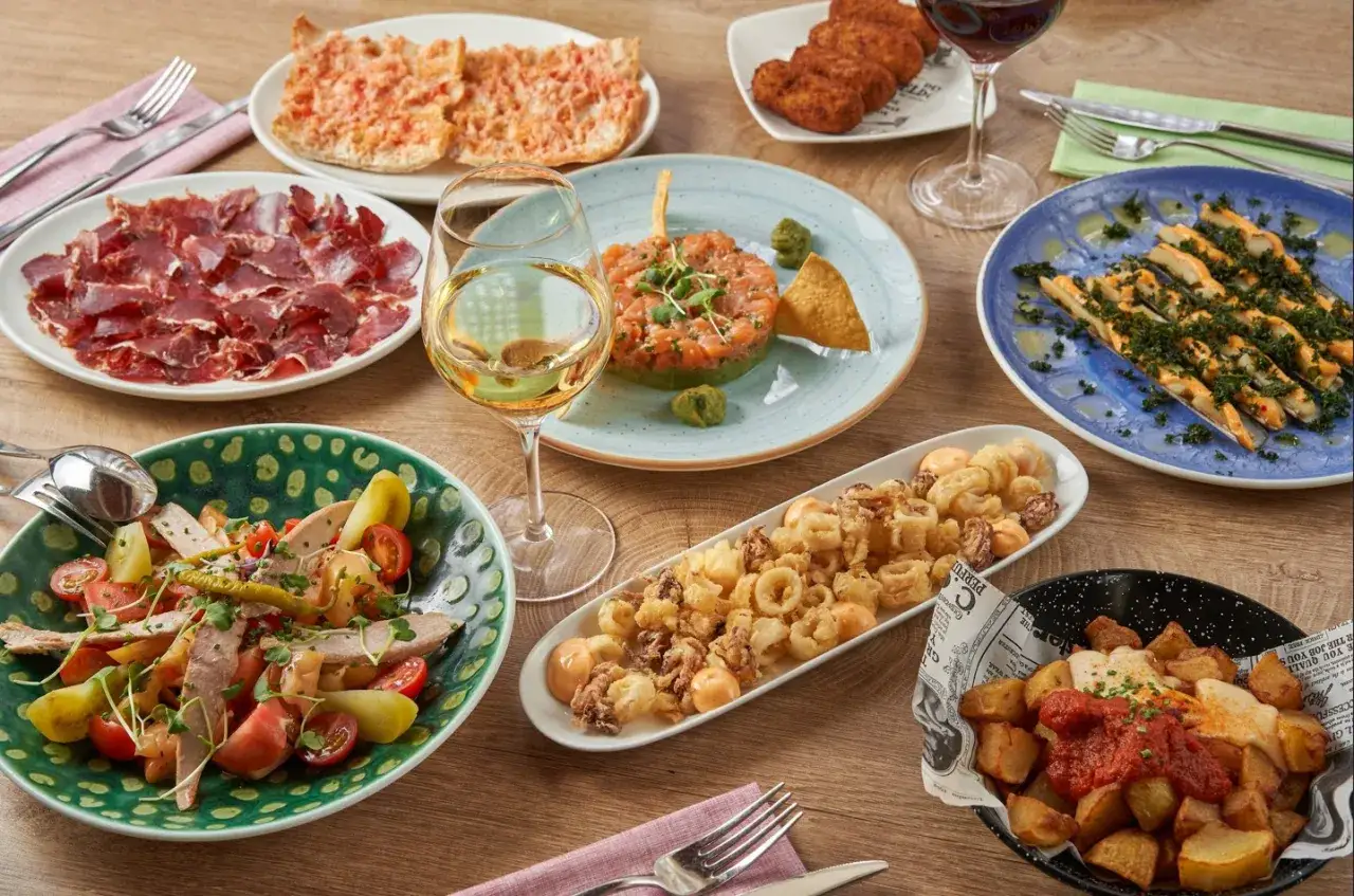 Una mesa llena de tapas deliciosas, ideal para decidir d&oacute;nde comer en Barcelona. Incluye pan con tomate, jam&oacute;n, ensalada, tartar de salm&oacute;n, calamares fritos y patatas bravas.