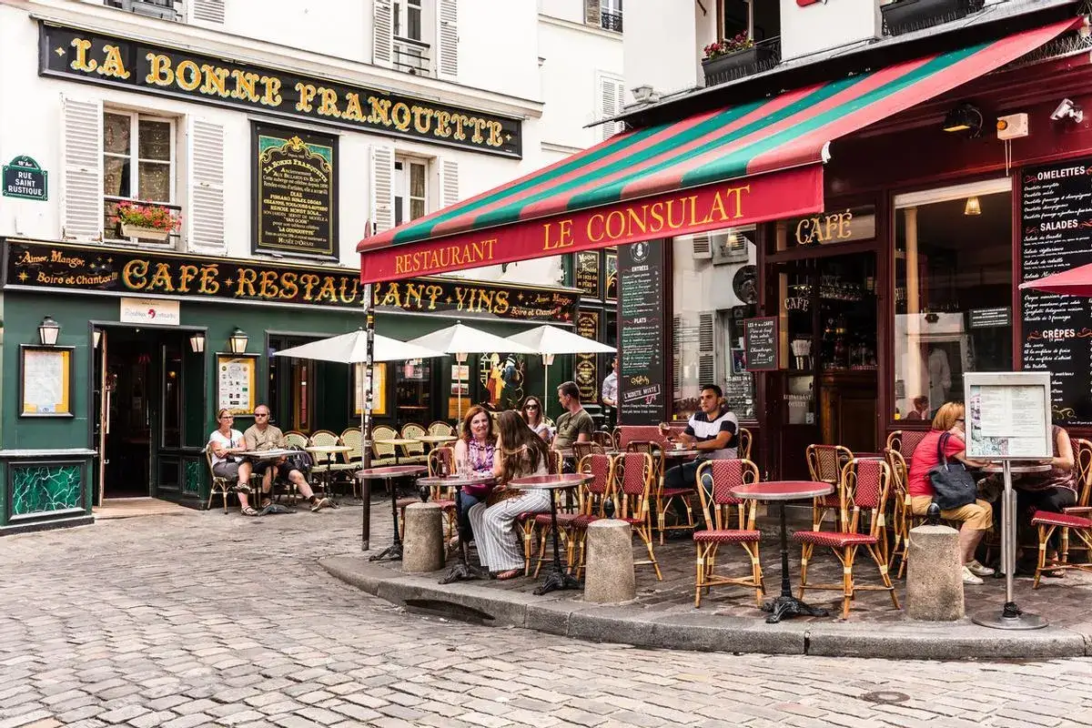 Caf&eacute;s y restaurantes en una calle adoquinada de Par&iacute;s, perfectos para saber d&oacute;nde comer en Par&iacute;s. Gente sentada en mesas al aire libre.