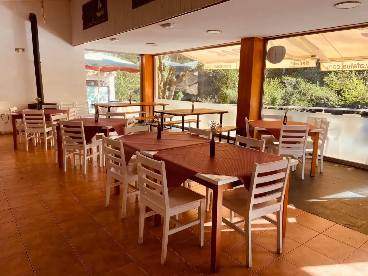 Mesas puestas con manteles rojos y sillas blancas, listas para comer en Pontedeume. Ventanales con vistas a la naturaleza.