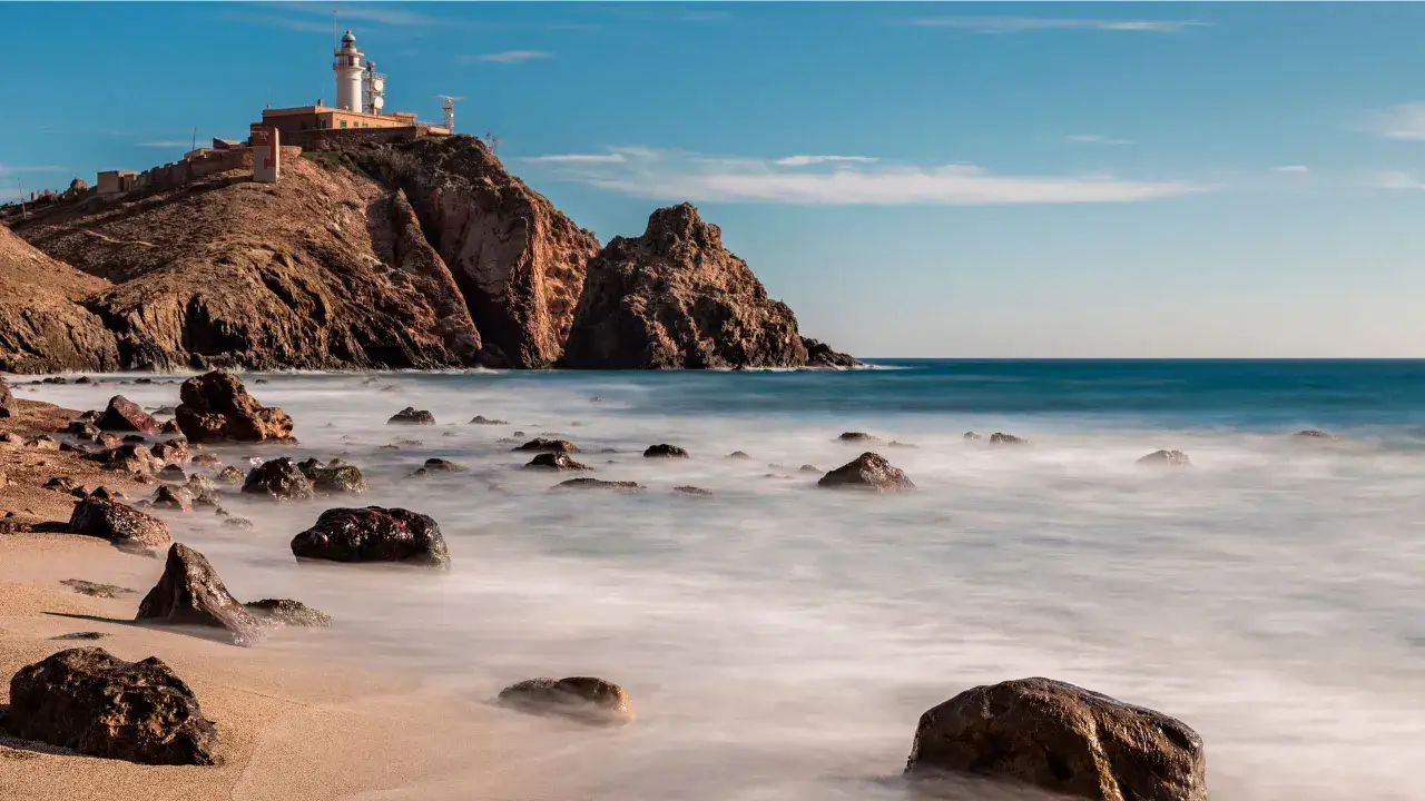 Faro en un acantilado rocoso sobre el mar, con rocas en la playa. Un lugar id&iacute;lico que ver en Cabo de Gata.