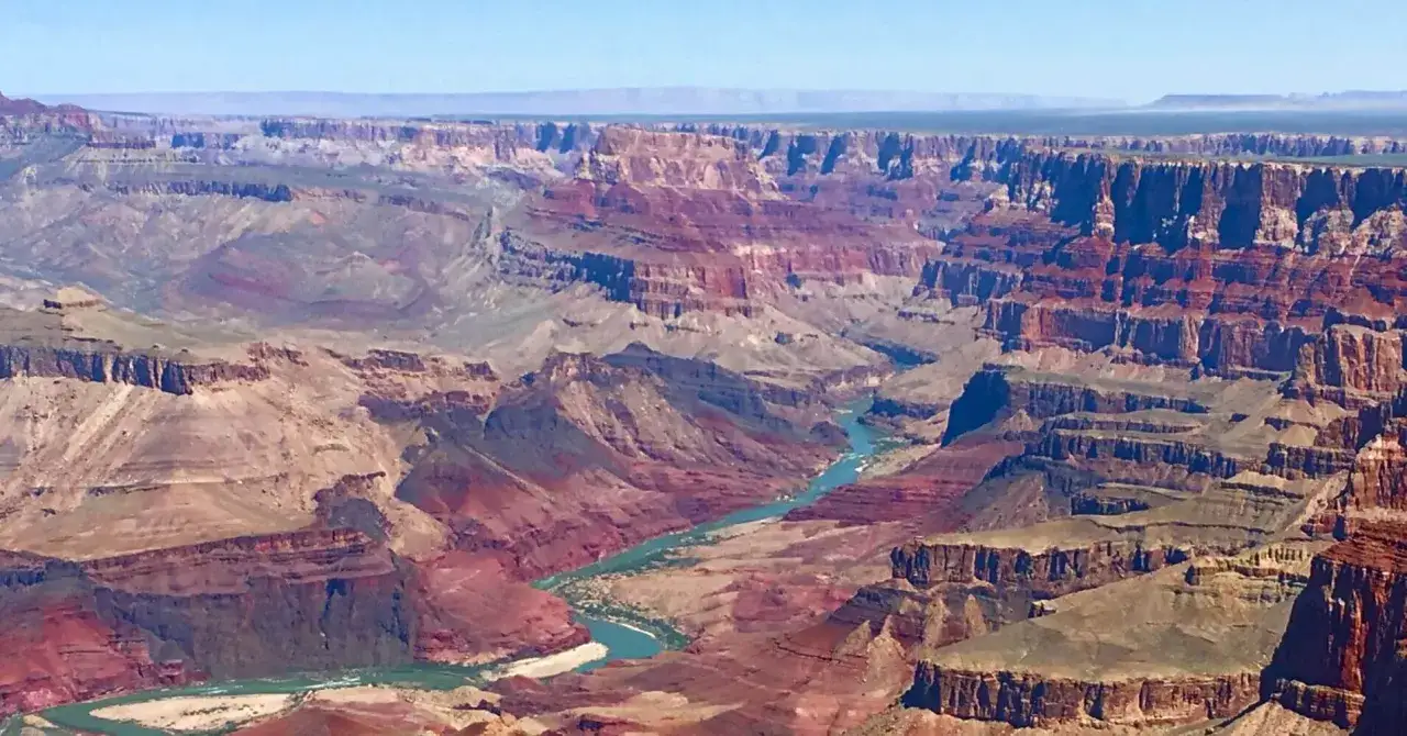 El majestuoso Cañón del Colorado, con sus capas rocosas de tonos rojizos y ocres, serpentea junto a un río turquesa bajo un cielo azul.