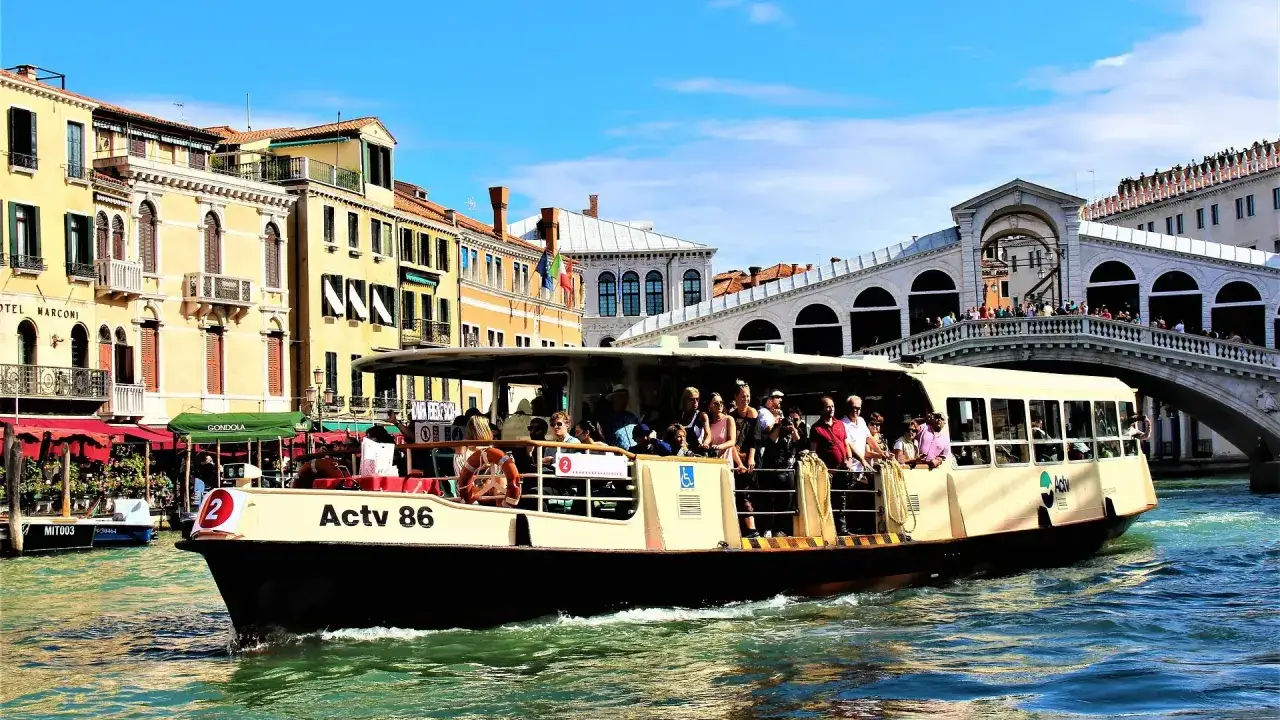 Barco lleno de turistas navega por el Gran Canal de Venecia, pasando bajo el Puente de Rialto. Un d&iacute;a perfecto para explorar la Venezia unica city pass.