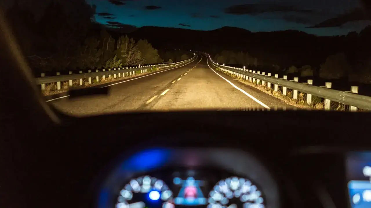 Conduciendo de noche, las luces de carretera iluminan el camino sinuoso y la barrera de seguridad. El tablero del coche se ve borroso.