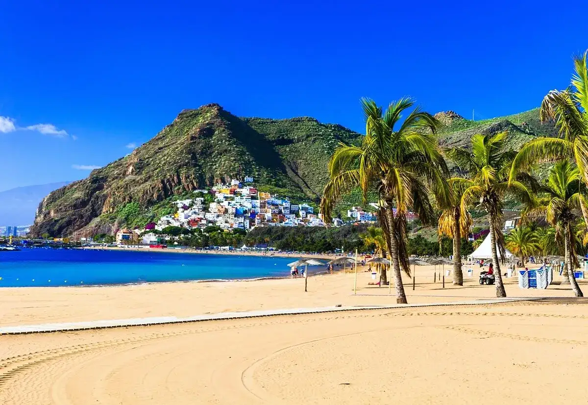 Playa de arena dorada con palmeras y un pueblo colorido en la ladera de una monta&ntilde;a. &iexcl;Un destino ideal donde viajar en agosto barato!