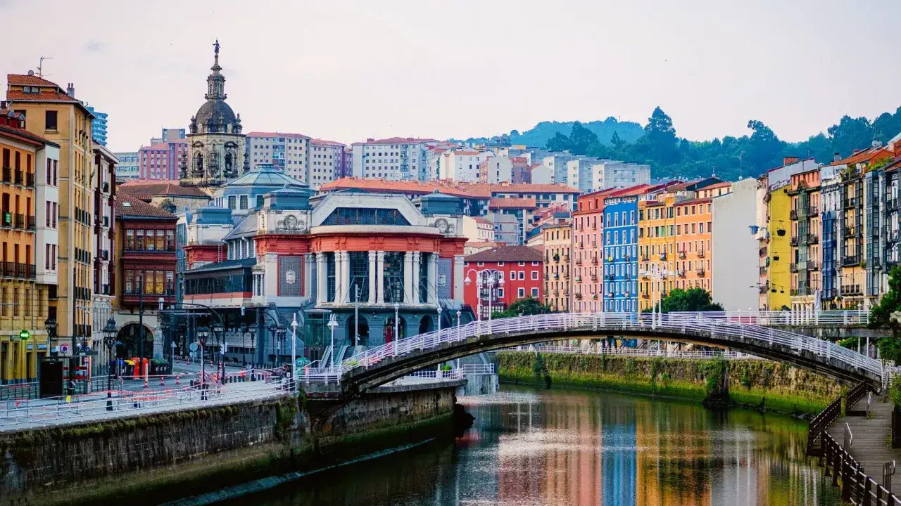 Un puente blanco cruza un r&iacute;o reflejando coloridas fachadas. Ideal para saber qu&eacute; ver en Bilbao en 2 d&iacute;as.