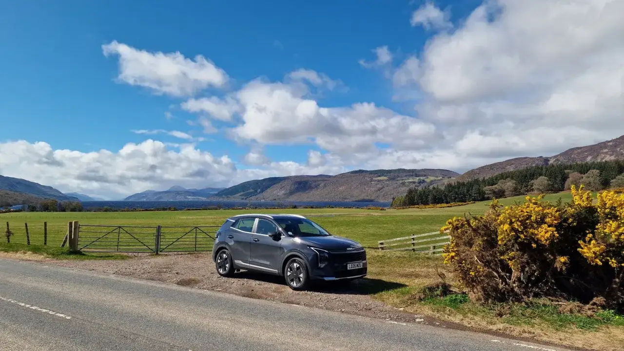 Coche gris aparcado junto a un lago escoc&eacute;s, ideal para viajar a Escocia por libre. Paisaje monta&ntilde;oso y cielo azul con nubes.